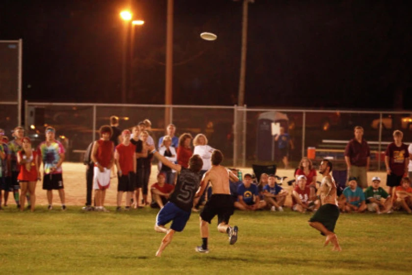 Players leaping for a disc under the stadium lights while a crowd watches from the sideline